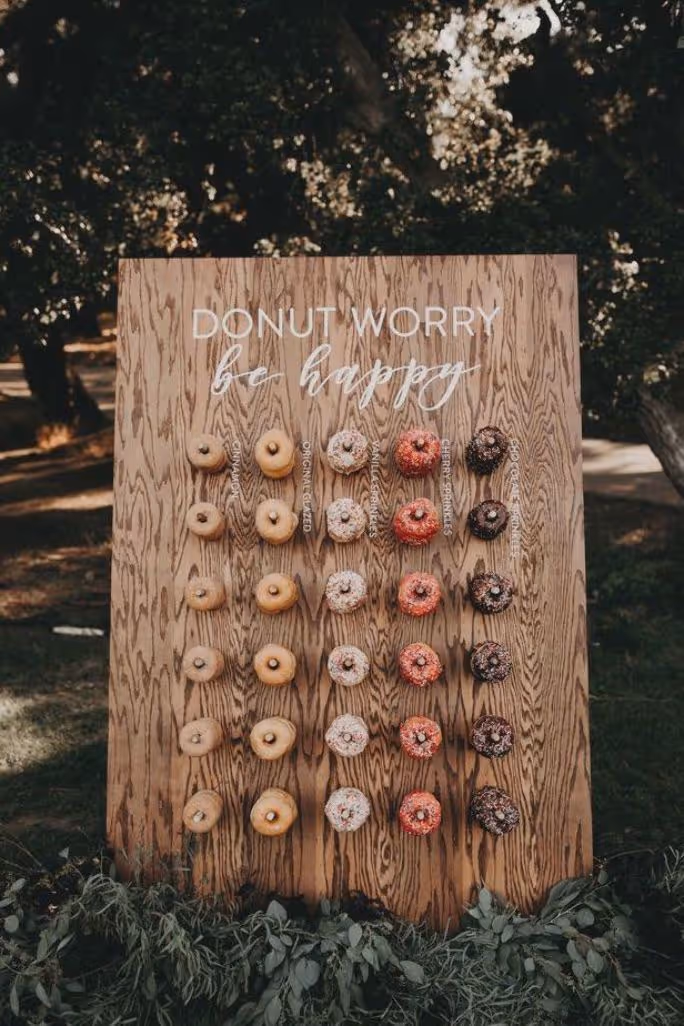Houten bord met donuts in vijf rijen en kolommen, erboven de tekst 'Donut worry be happy'.