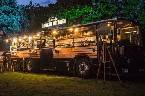Een verlicht eetkraam in een omgebouwde bus met het bord 'Drive by Burger Kitchen' 's avonds buiten tussen bomen.