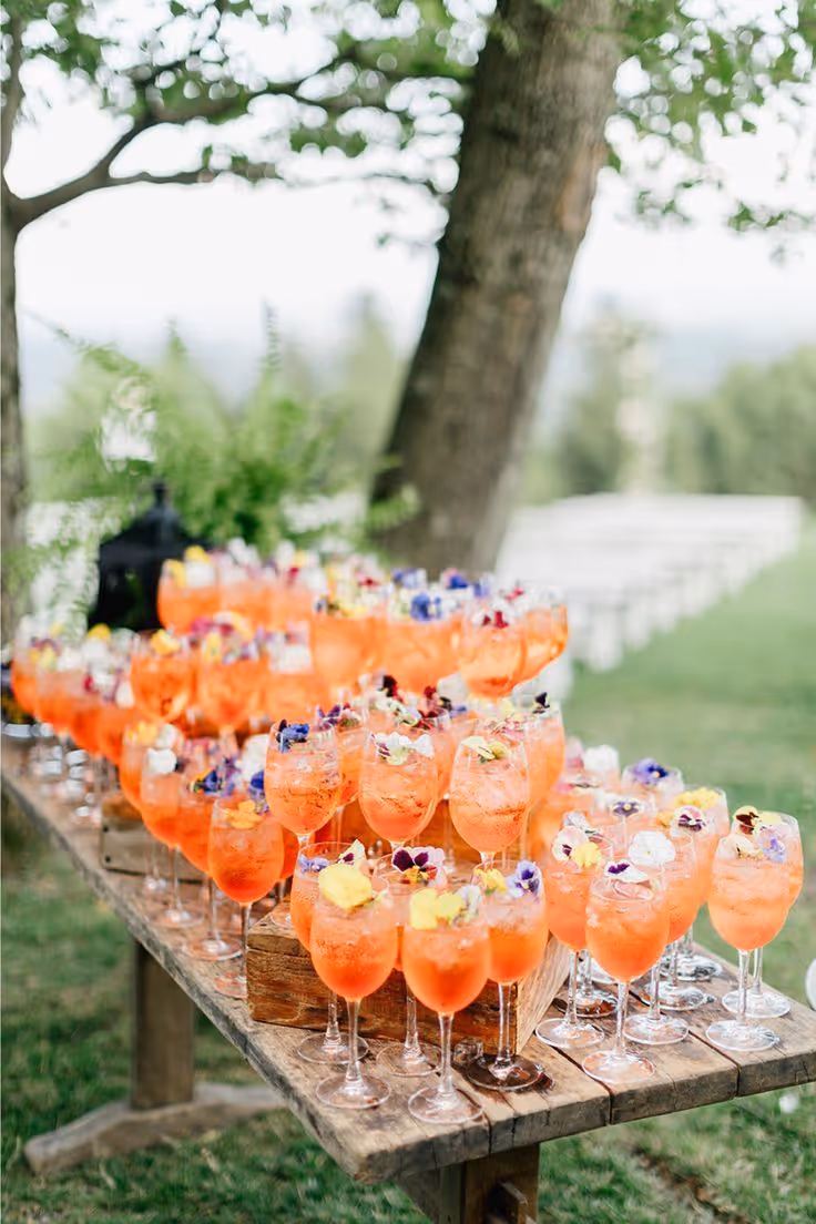 Tafel buiten met rijen glas met oranje aperitiefdrankjes versierd met eetbare bloemen.