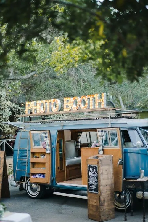 Blauwe vintage bus met open schuifdeuren en verlichte letters 'PHOTO BOOTH' op het dak, gebruikt als fotohokje in een buitenomgeving met bomen.
