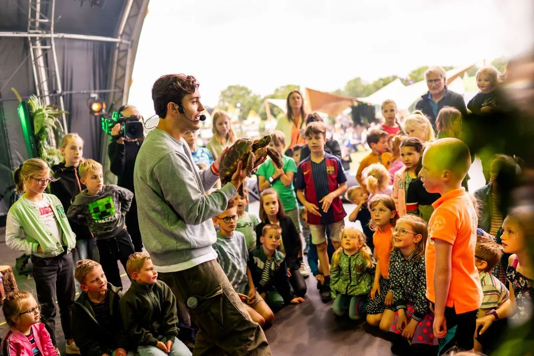 Man met headset toont een schildpad aan een groep nieuwsgierige kinderen tijdens een buitenactiviteit.