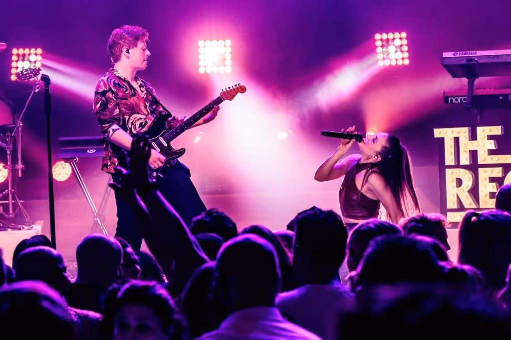 Een gitarist en zangeres die live optreden op een podium met paars en roze verlichting en een publiek op de voorgrond.