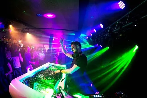 DJ draait muziek achter een mengpaneel tijdens een feest met kleurrijke verlichting en dansende menigte.