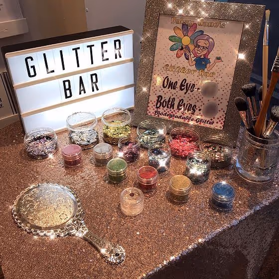 Glitter bar setup with small containers of various colored glitters, a vintage hand mirror, makeup brushes, and a lightbox sign reading 'GLITTER BAR' on a sparkly rose gold tablecloth.