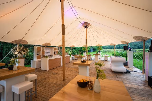 Strakke inrichting onder een grote witte tent met houten tafels, witte barkrukken en groene buitenomgeving.
