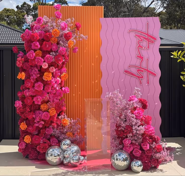 Roze en oranje bloemenarrangementen rondom een roestvrijstalen Plexiglas standaard met roze en oranje achtergrondpanelen en 'thirty' in roze neonletters.
