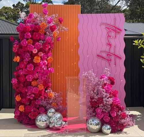 Felgekleurde bloemen in roze, oranje en roodarrangement rond een roze en oranje decoratiepaneel met zilveren discoballen op de grond.