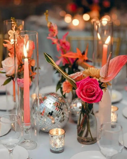Tafeldecoratie met brandende roze kaarsen in glazen houders, bloemen in vazen en een glitterende discobal op een feestelijke tafel.