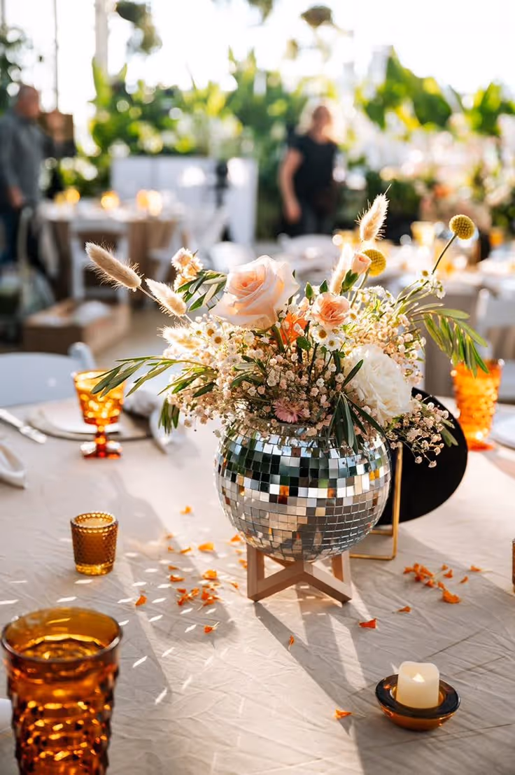 Tafel met een glinsterende disco-balvaasje gevuld met diverse pastelroze bloemen en groene bladeren als tafeldecoratie.