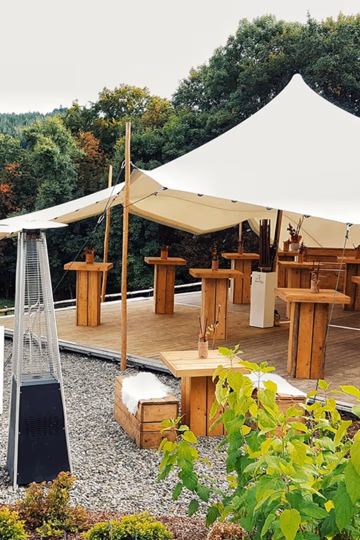 Buitenlounge met houten tafels en banken onder een witte tent, omgeven door groen en een buitenverwarmer.