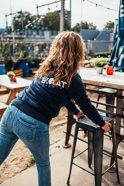 Vrouw met golvend haar in een blauwe trui en spijkerbroek zet een kruk bij een houten tafel in een buitenruimte.
