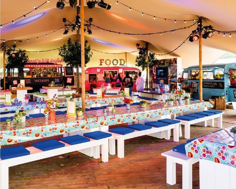 Groot feesttent met lange tafels bedekt met bloemrijke tafelkleden en blauwe kussens op witte banken, met foodtrucks en sfeerverlichting.
