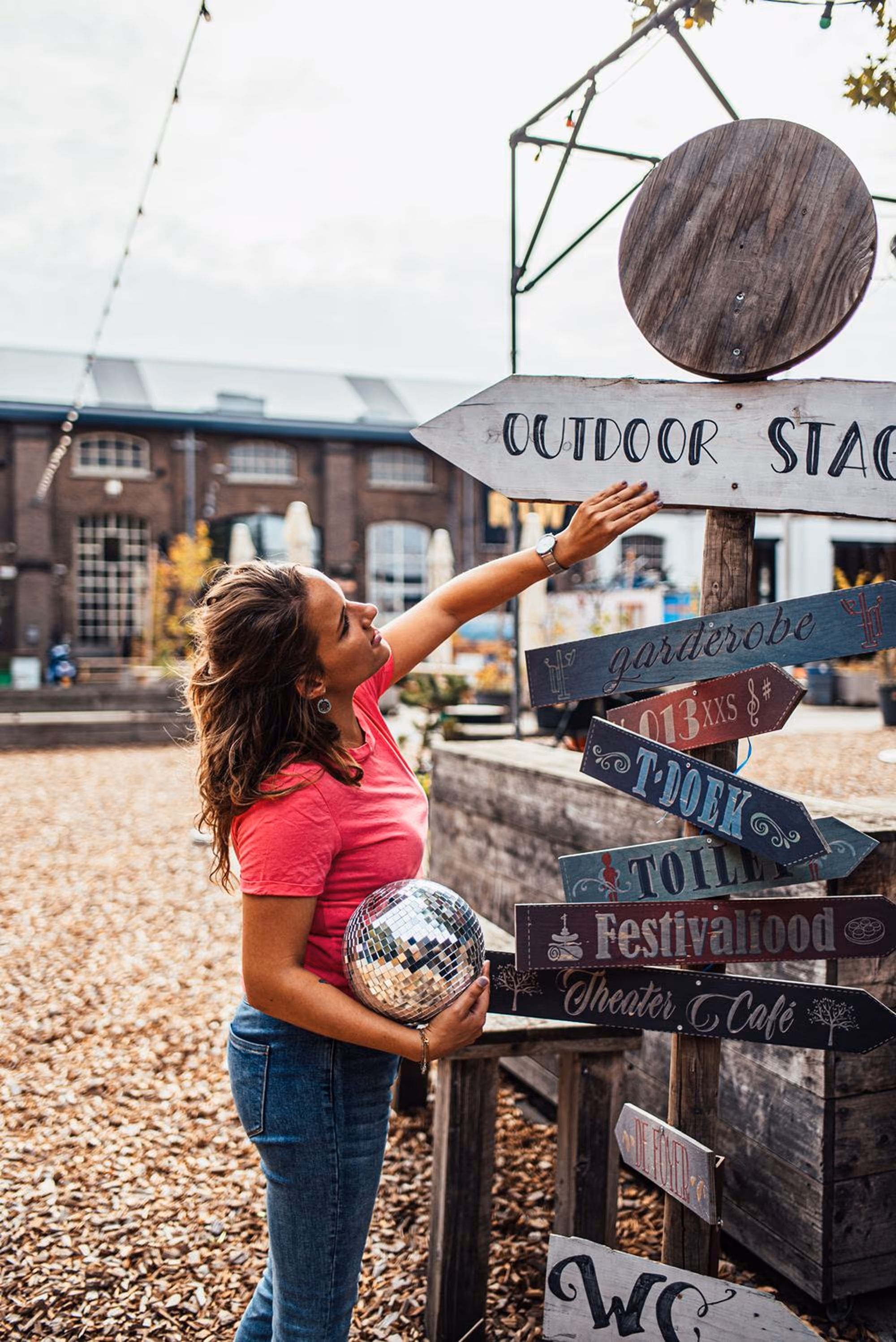 Vrouw in een roze shirt en jeans houdt een discobal en wijst naar een houten bord met richtingen bij een buitenlocatie.