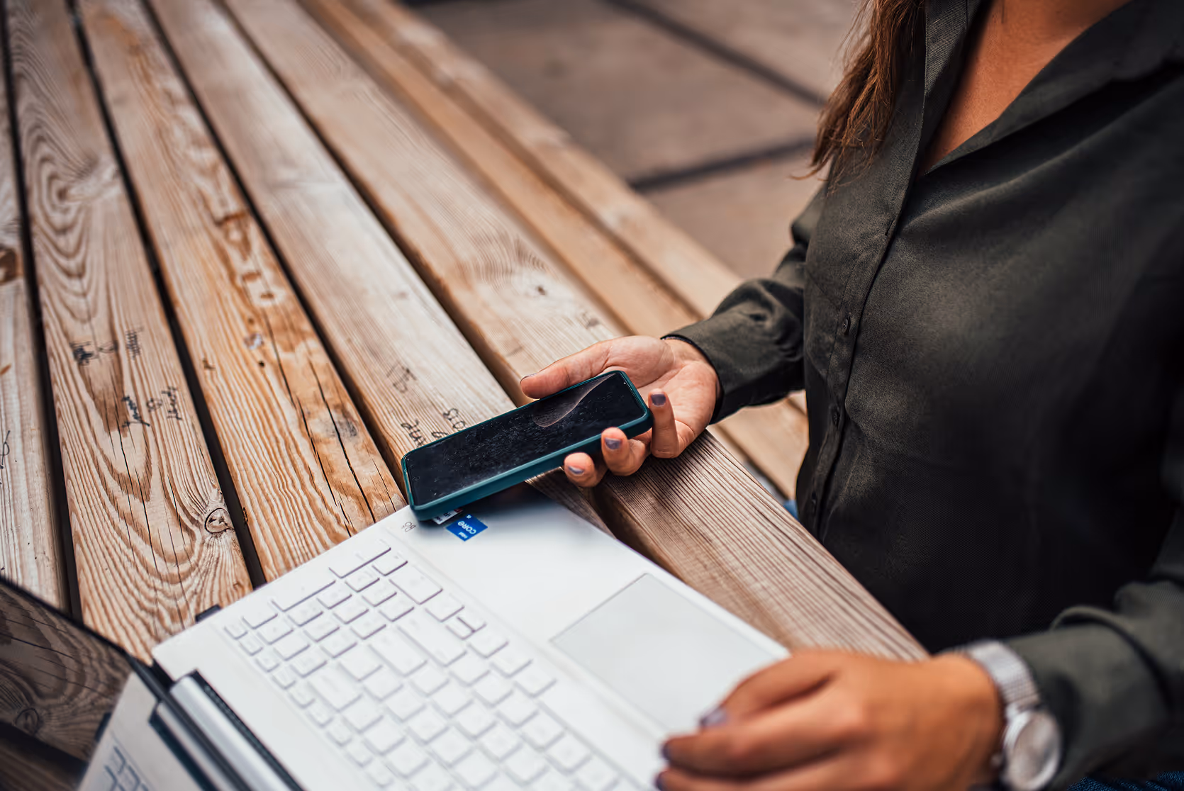 Karin met donker overhemd houdt een smartphone vast boven een witte laptop op een houten tafel.