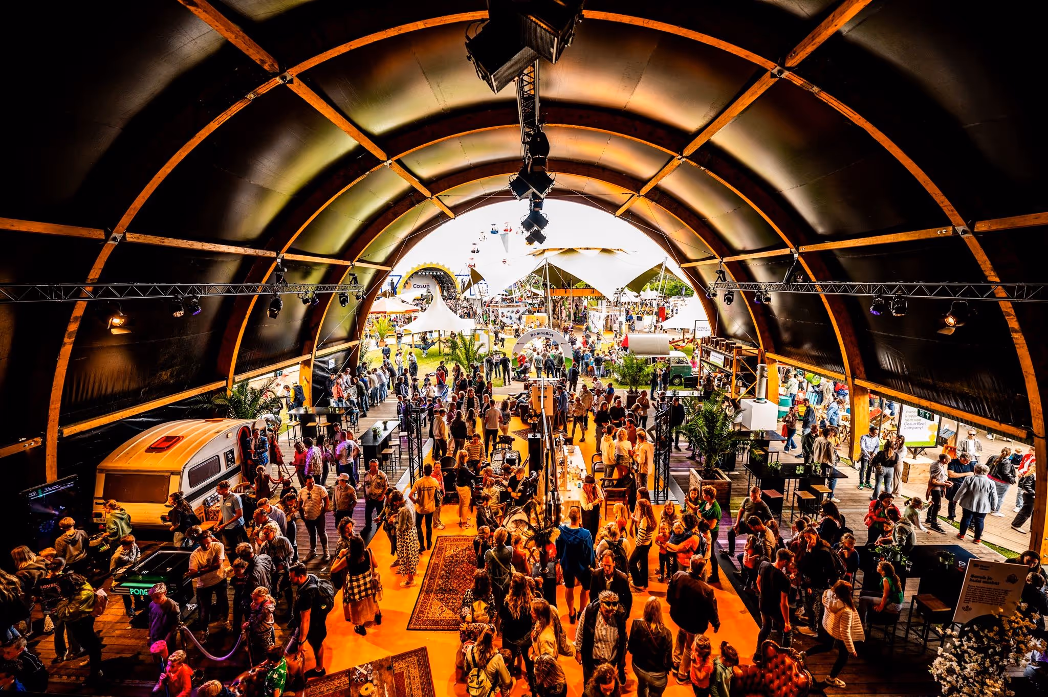 Drukke beurs of markt met veel mensen onder een grote overdekte hal, met stands en tafels, en zicht op een buitengebied met meer bezoekers.