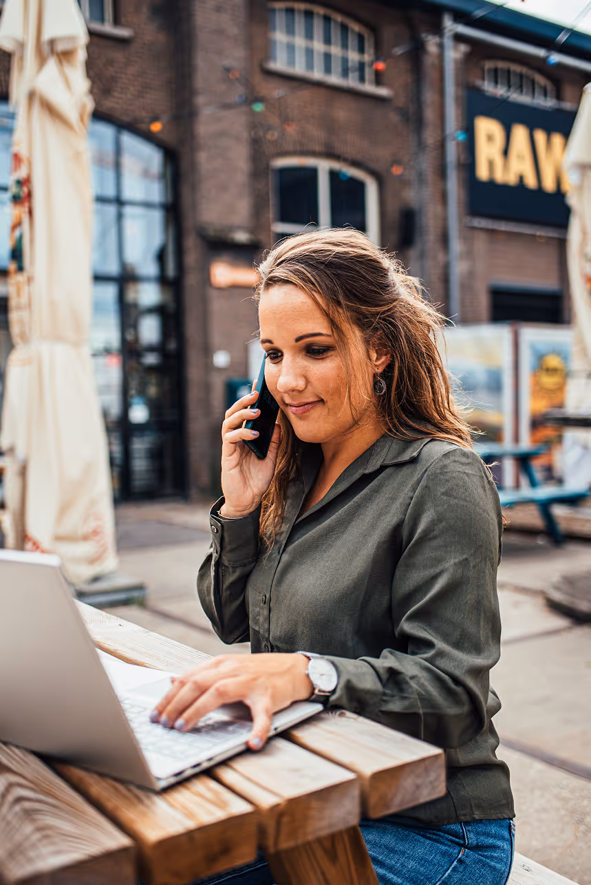 Karin die buiten aan een houten tafel werkt op een laptop en op haar telefoon praat.