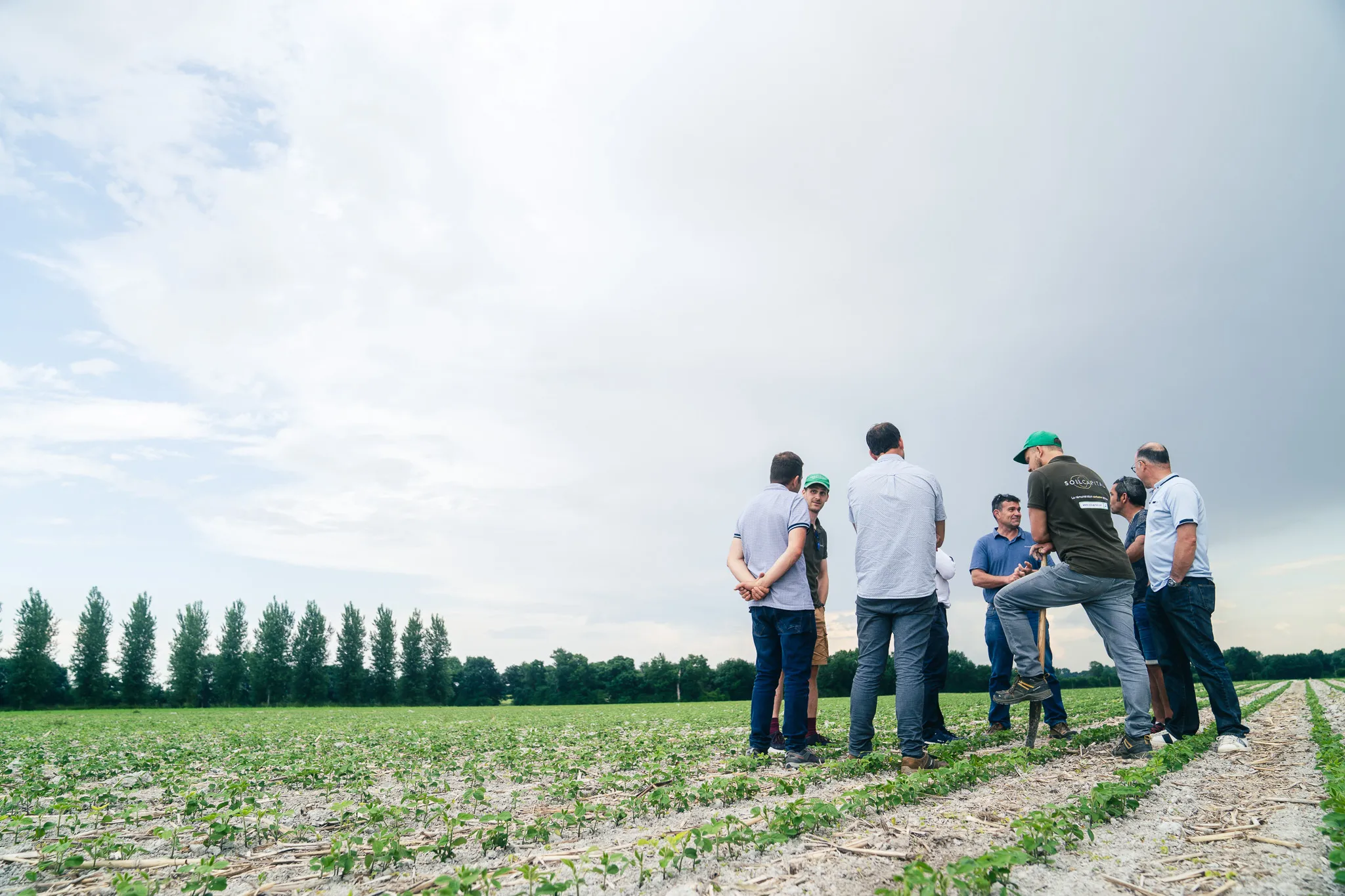Group of people standing and talking in a field with young crops under a cloudy sky.