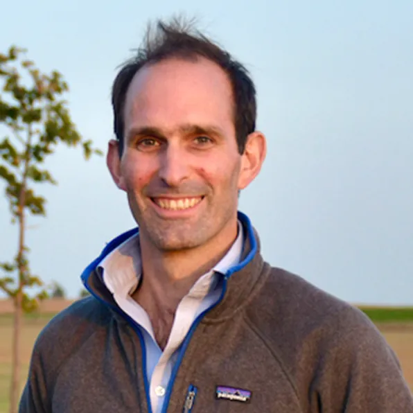 Smiling man with short dark hair wearing a gray fleece jacket outdoors with a tree and open field in the background.