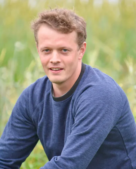 Young man with short curly hair wearing a blue sweatshirt outdoors with greenery in the background.