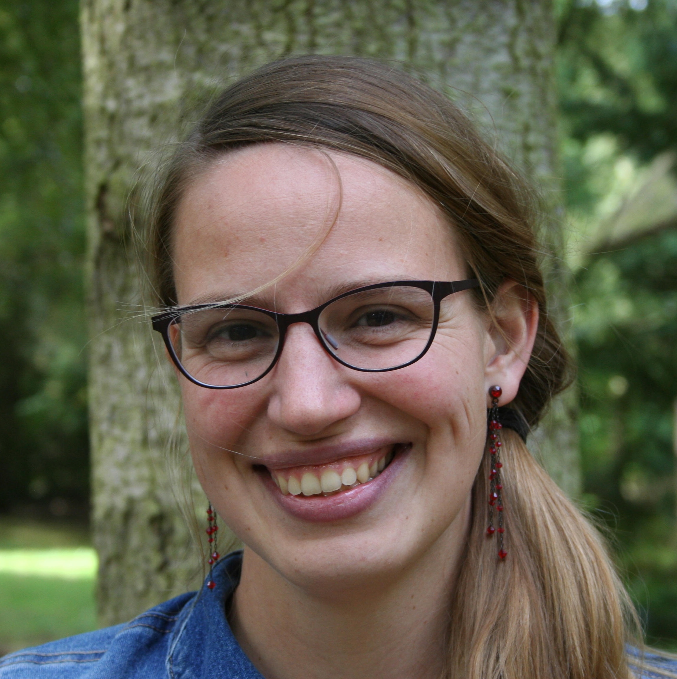 Smiling woman with glasses, long light brown hair tied back, and red bead earrings in an outdoor setting with trees.