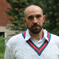 Man with a bald head and beard wearing a white cable-knit sweater with blue and red trim, standing outdoors with trees and a brick building in the background.