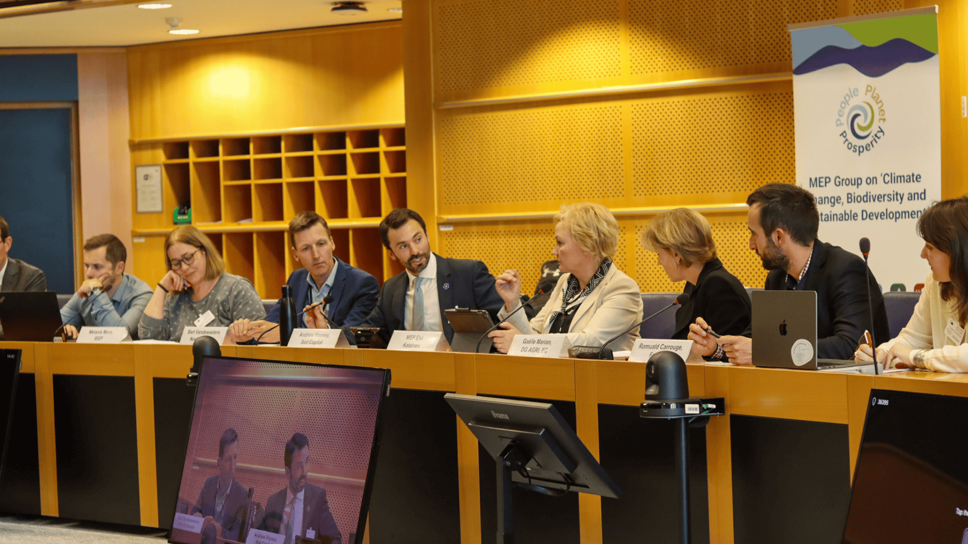 A panel of eight people engaged in discussion at a conference table with nameplates and microphones, and a banner in the background about climate change, biodiversity, and sustainable development.