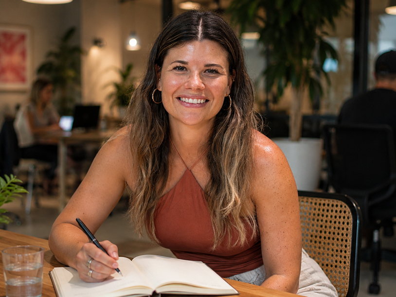 Smiling woman with long hair writing in an open notebook at a wooden table in a cozy workspace.