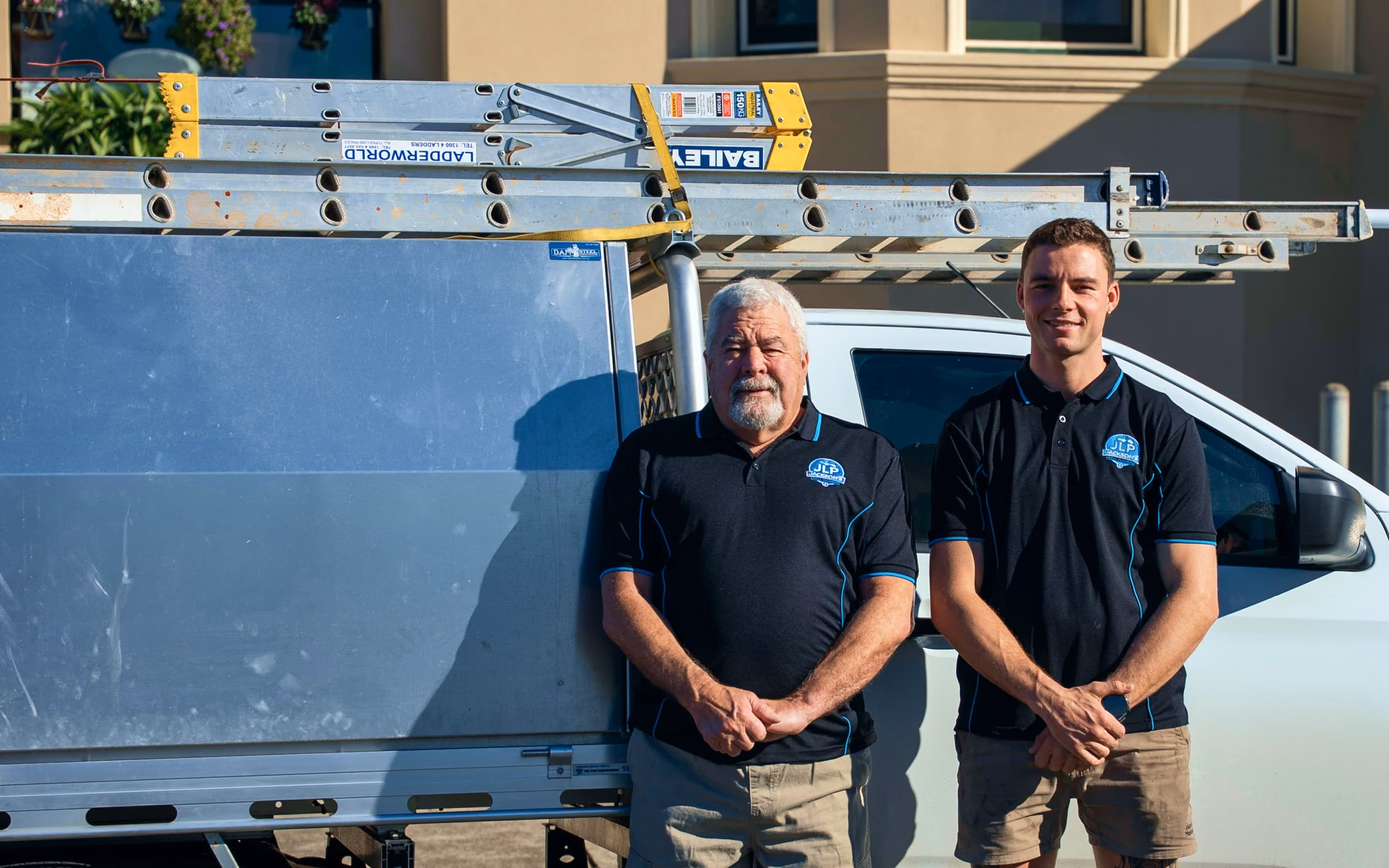 Two men in black company shirts standing in front of a white work vehicle with ladders.