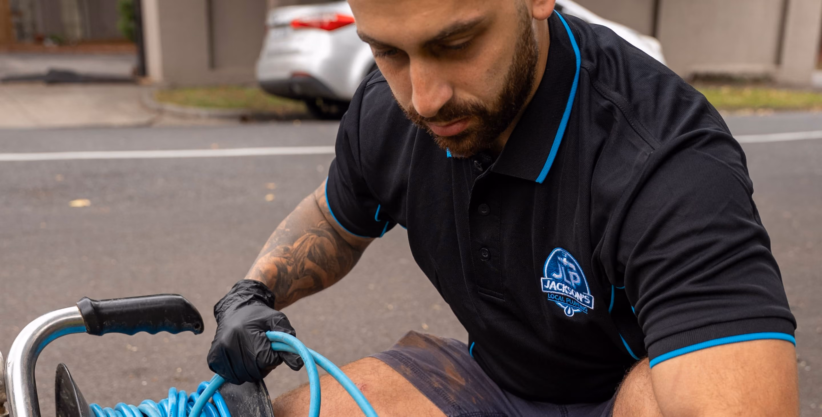 Man in black gloves coiling a blue hose, wearing a Jackson's Local Plumbing shirt.