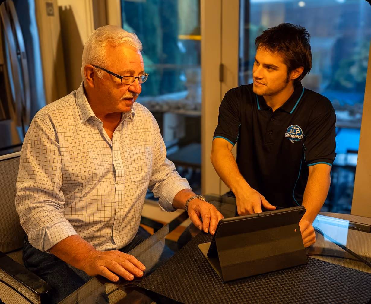 Young man in a Jackson's shirt showing tablet content to an older man in glasses at a glass table.