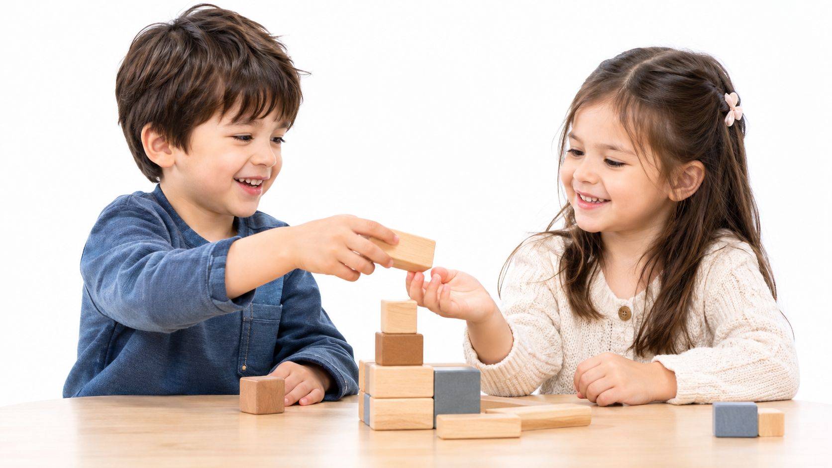 A young boy and girl sitting at a wooden table smiling while building with wooden blocks.