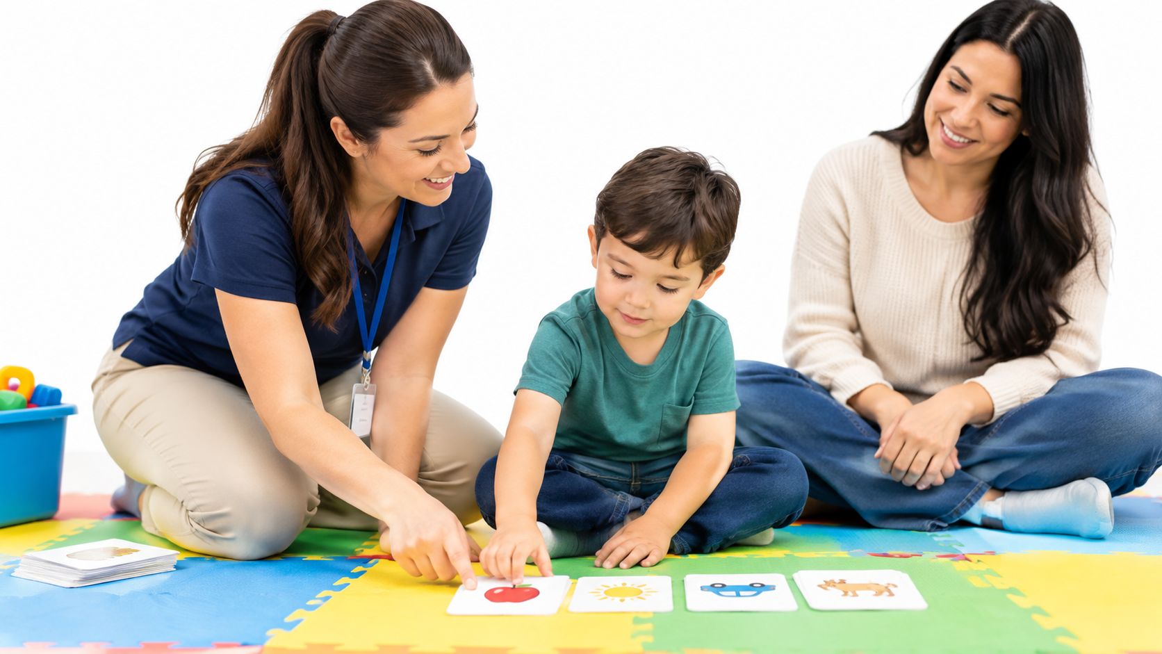 An ABA therapist and a mother help a young boy learn using educational cards on a colorful mat.