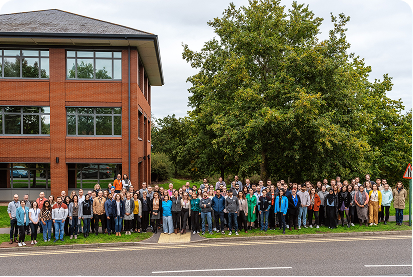 Large group of people standing outdoors near a brick building and large tree on a cloudy day.