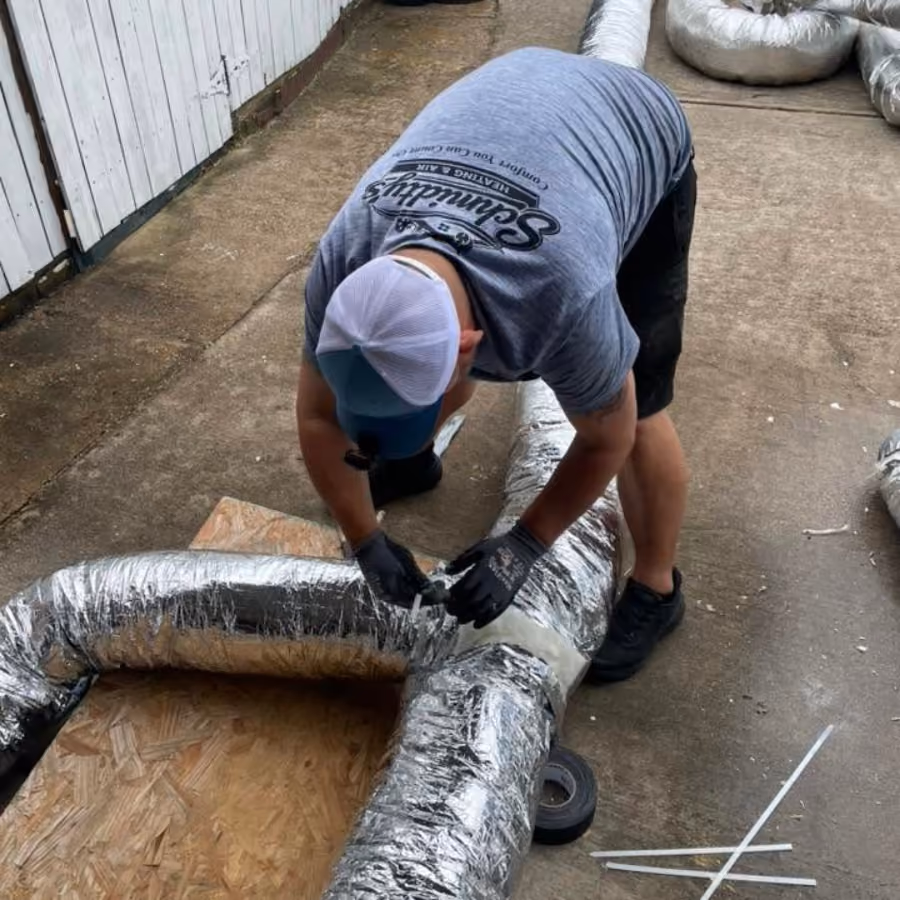 Man in a cap and gloves working on silver insulated ducting laid on concrete floor outdoors.