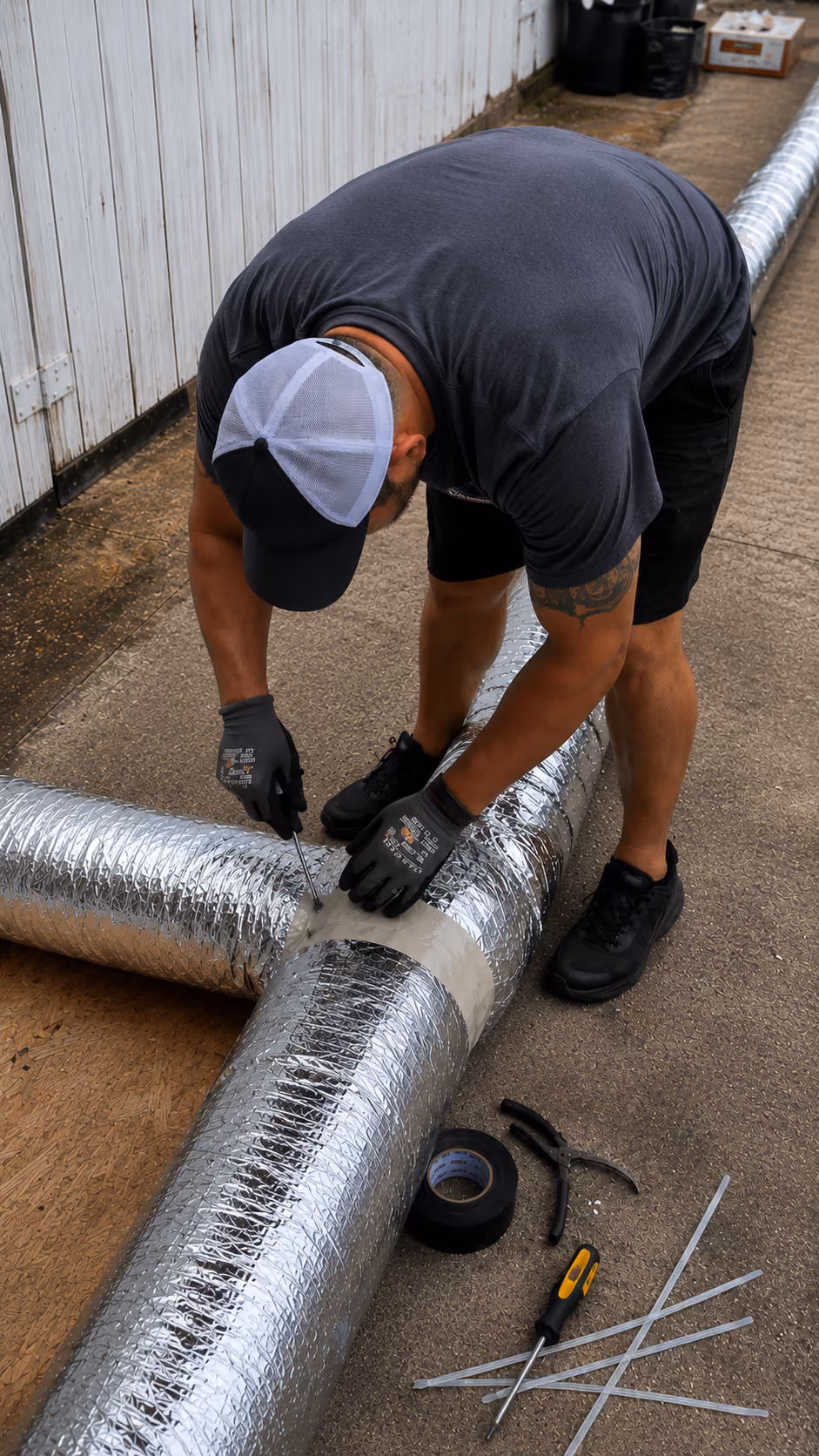 Man wearing gloves and cap working on connecting insulated HVAC ducts with tape and tools on concrete floor.