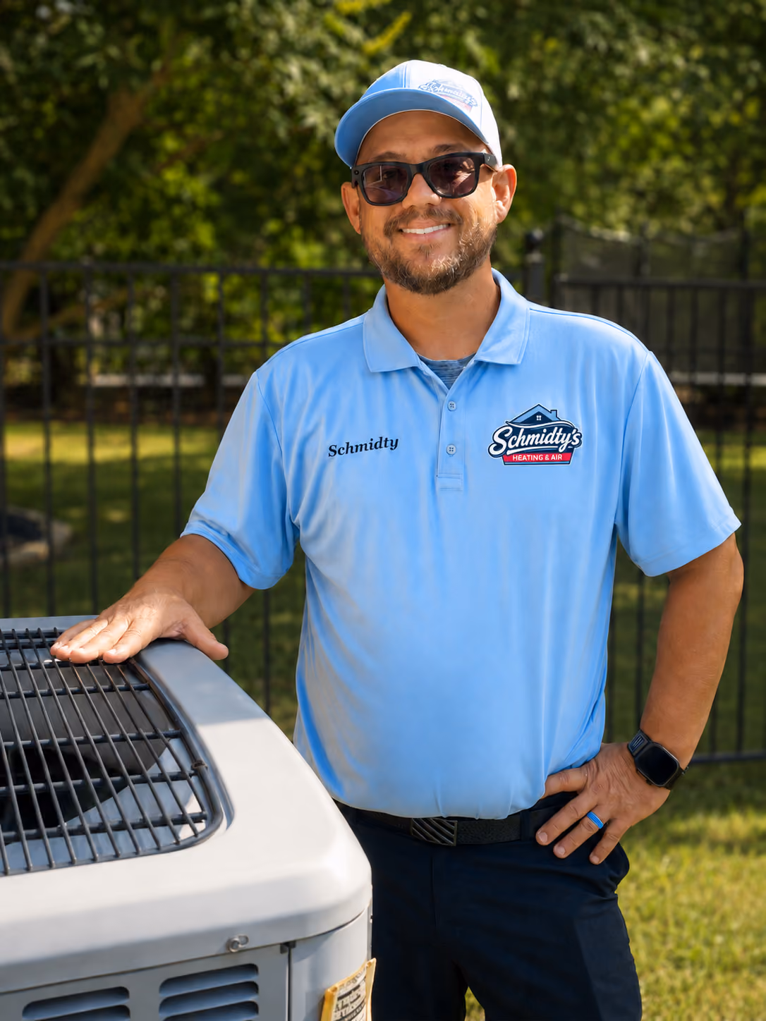 Man in blue Schmidty's Heating & Air uniform standing outdoors by an HVAC unit, smiling.