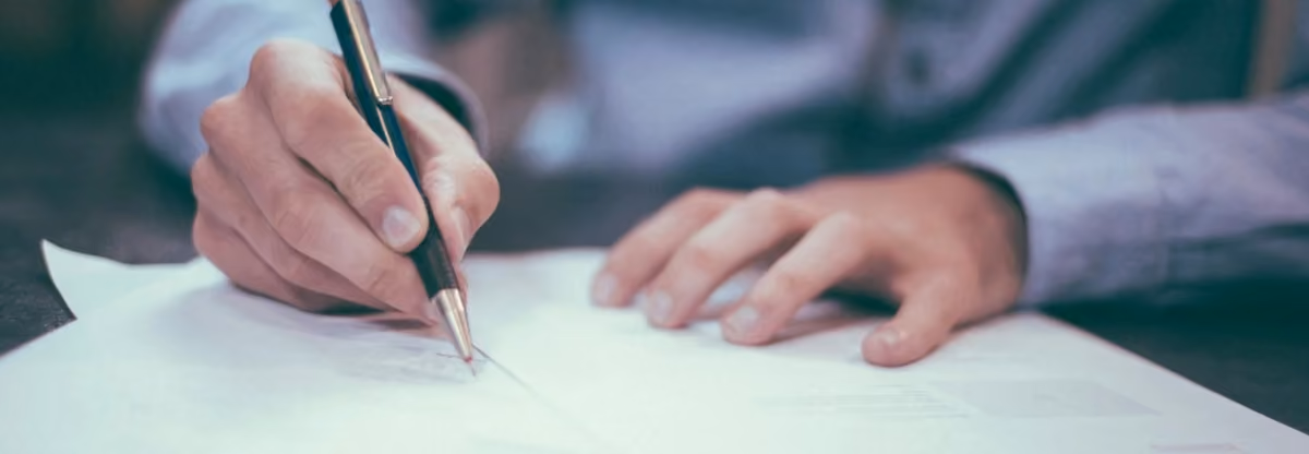 Close-up of a person writing on a white sheet of paper with a pen on a dark surface.