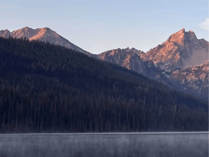 Mountain peaks lit by sunset above a dark forest with mist over a calm lake.
