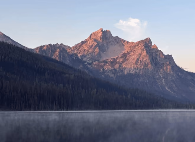 Mountain range lit by sunset light behind forest and mist over calm lake water.