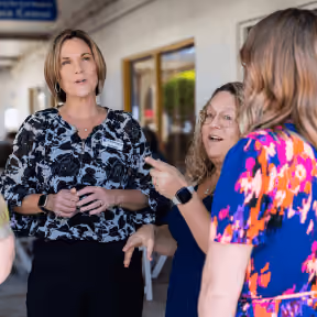Three women in conversation outside a building, one speaking and others listening attentively.
