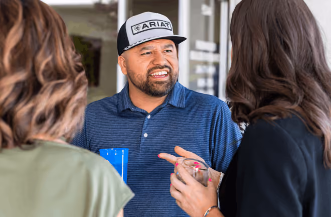 Man wearing an Ariat cap and blue shirt talking to two women holding drinks.