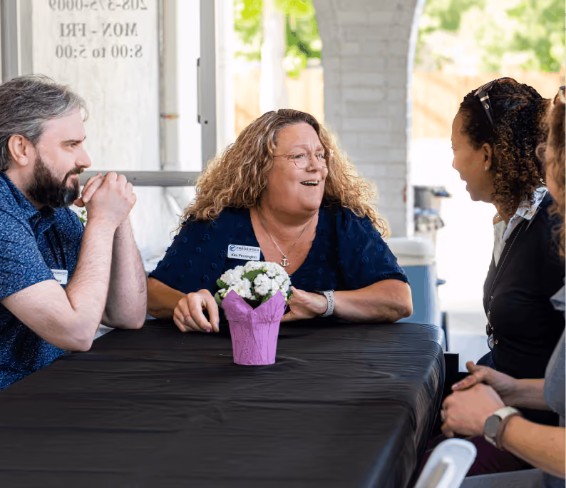 Four people sit around a table covered with a black cloth, talking and smiling around a flower pot.