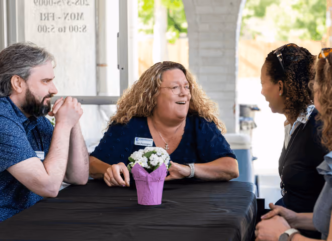 Three people sitting at a table talking with a potted flower in a purple wrapper in the center.