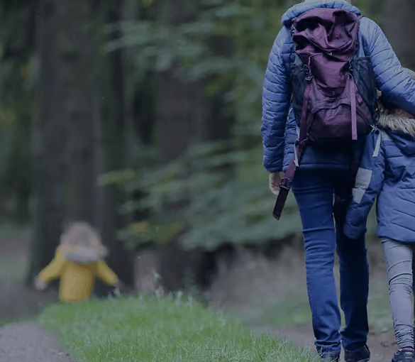 Person with purple backpack walks on forest path holding child's hand while another child runs ahead.
