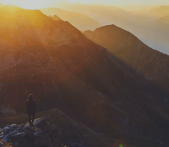 A person standing on a rocky peak watching a sunlit mountain range at dawn or dusk.