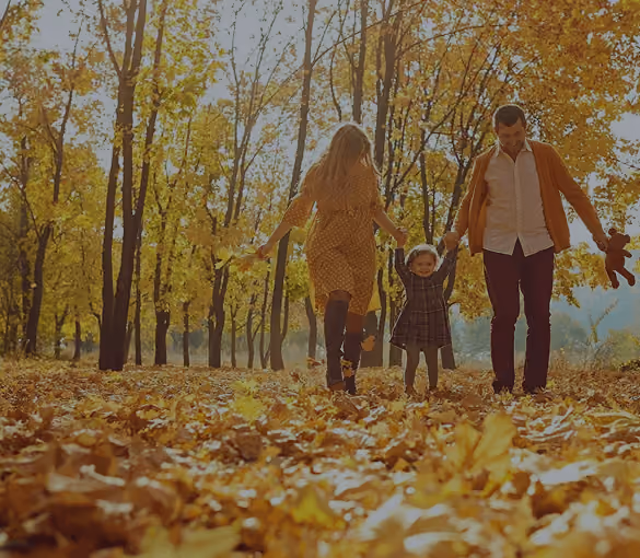 Parents holding hands with their child walking in a park covered with autumn leaves.