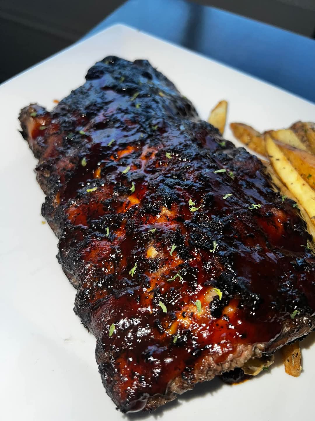 Barbecue ribs glazed with sauce and sprinkled with herbs served with potato wedges on a white plate.