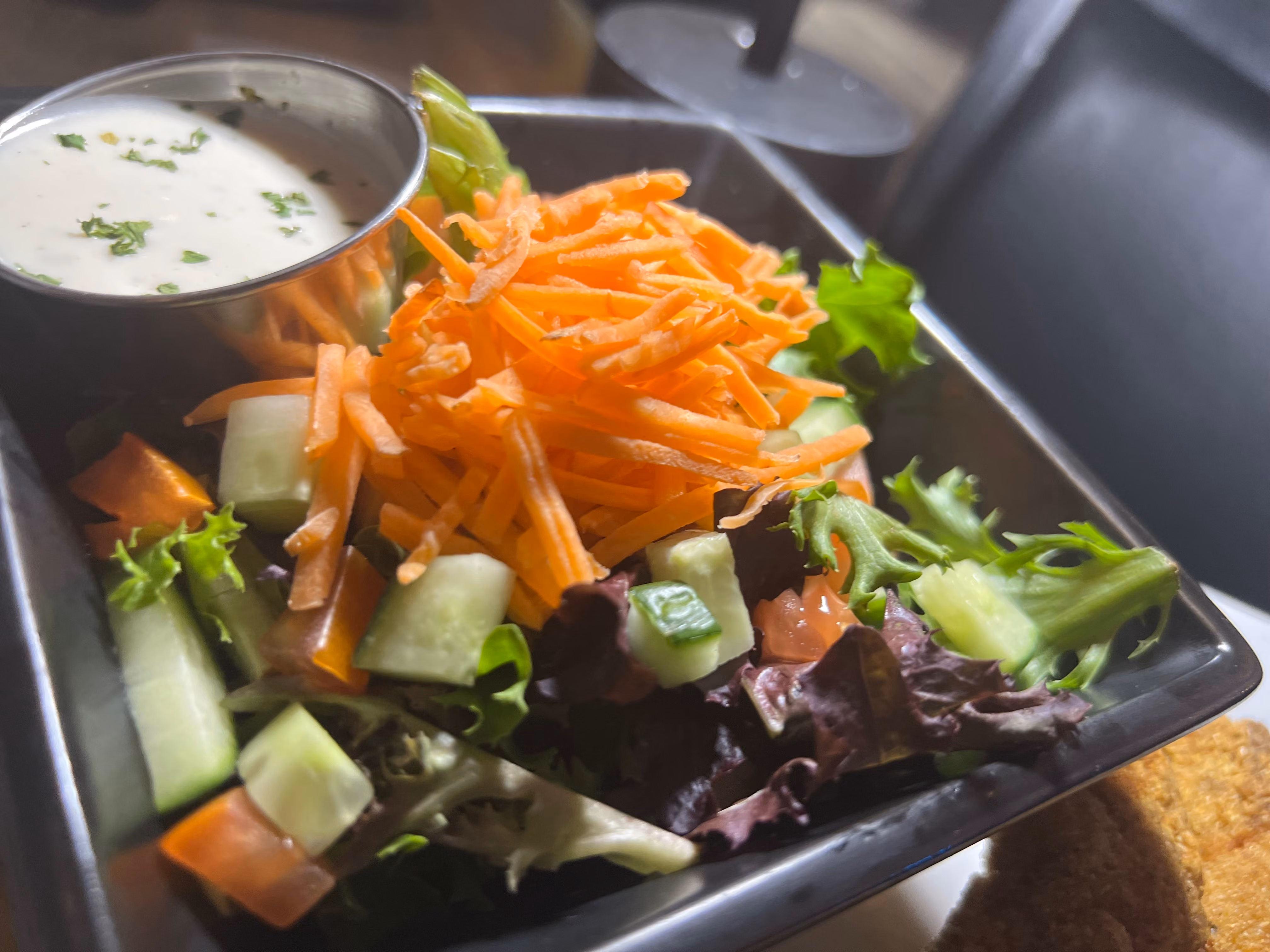 Fresh salad with shredded carrots, cucumber, lettuce, tomato, and a side of creamy dressing in a black bowl.