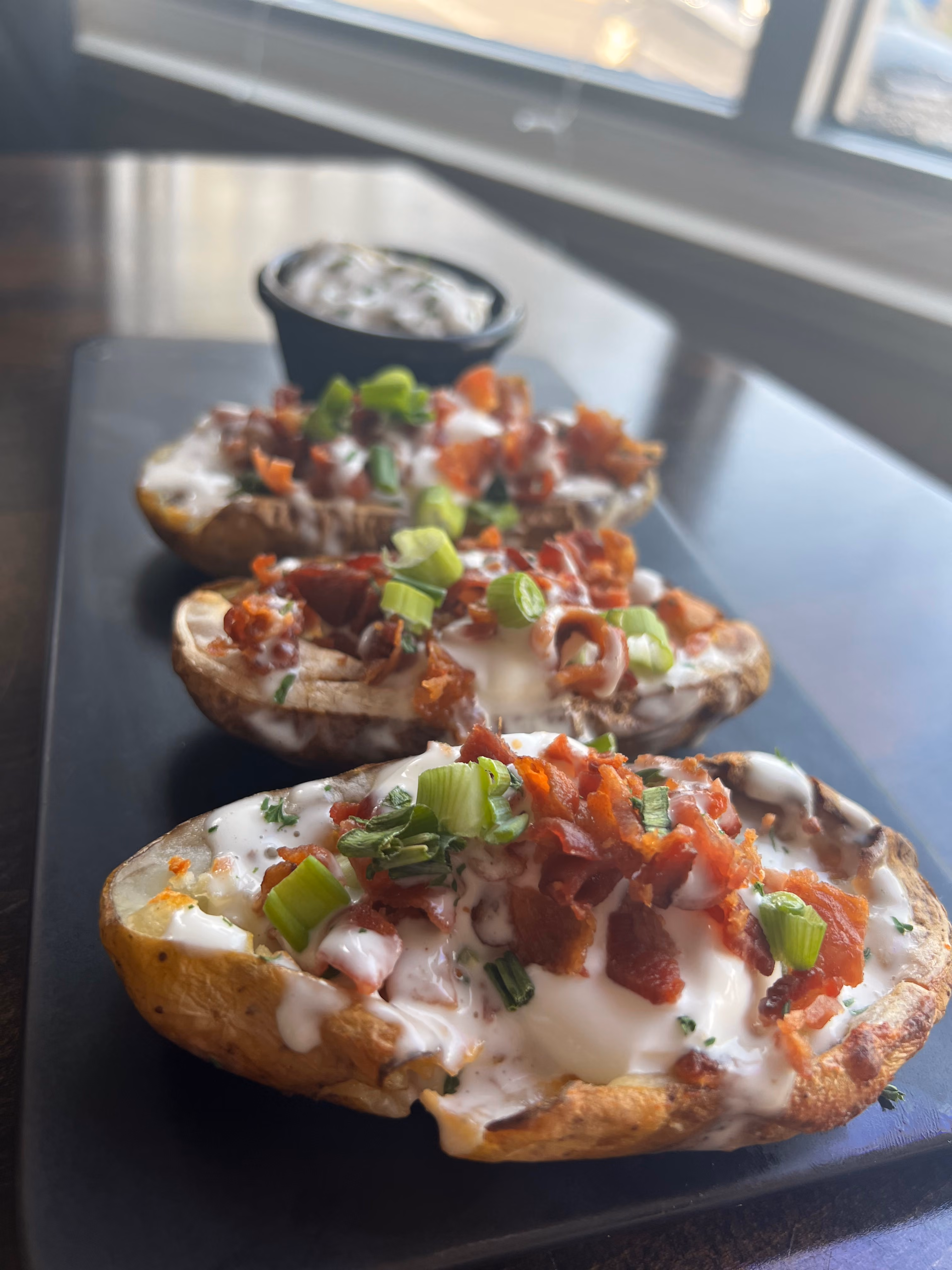 Three loaded potato skins with sour cream, bacon bits, and green onions on a black tray.