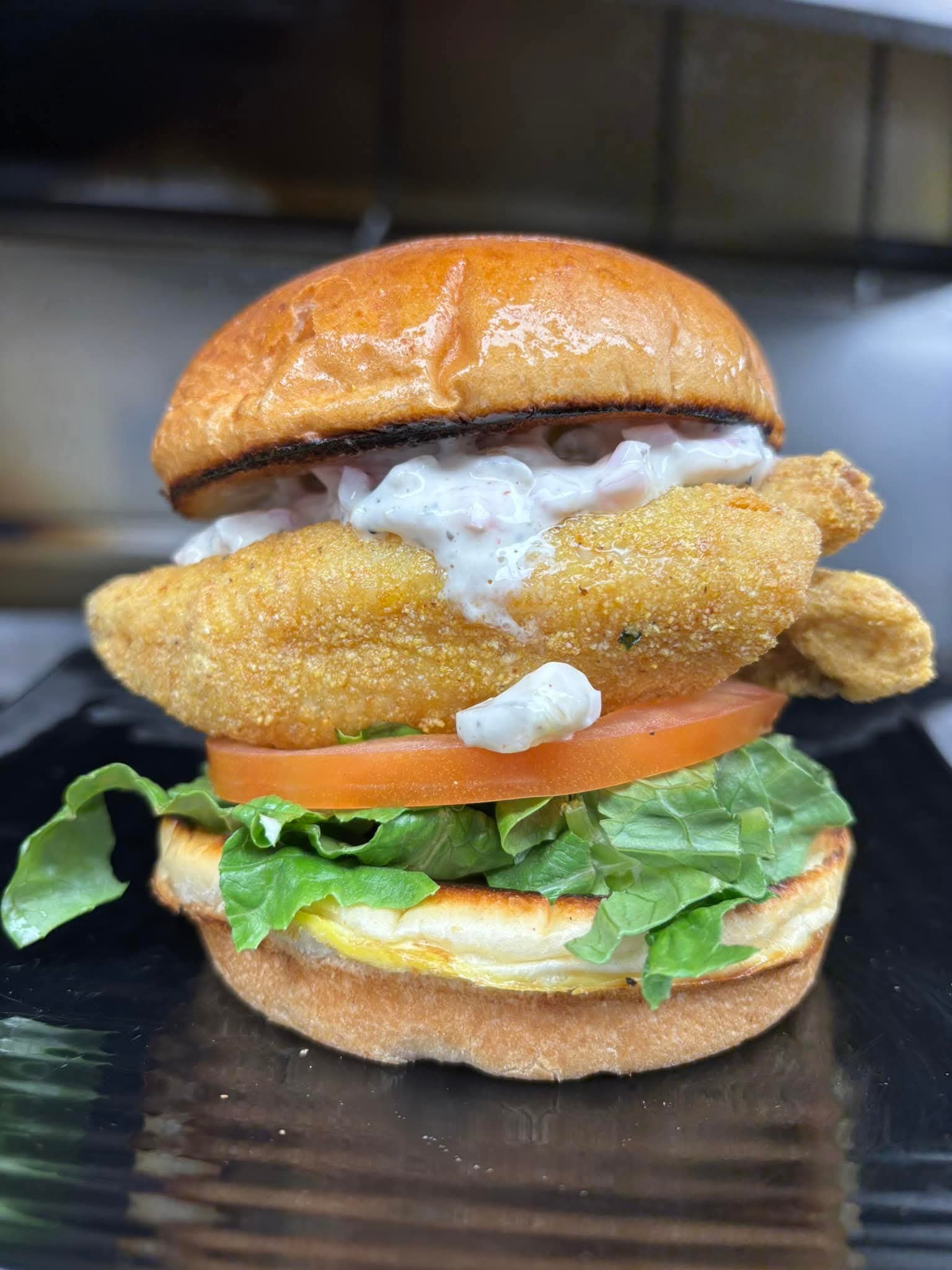 Fried fish sandwich with lettuce, tomato, tartar sauce, and toasted bun on a black plate.
